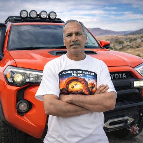Man standing in front of a red Toyota truck with 'Adventure Starts Here' t-shirt, desert landscape in the background