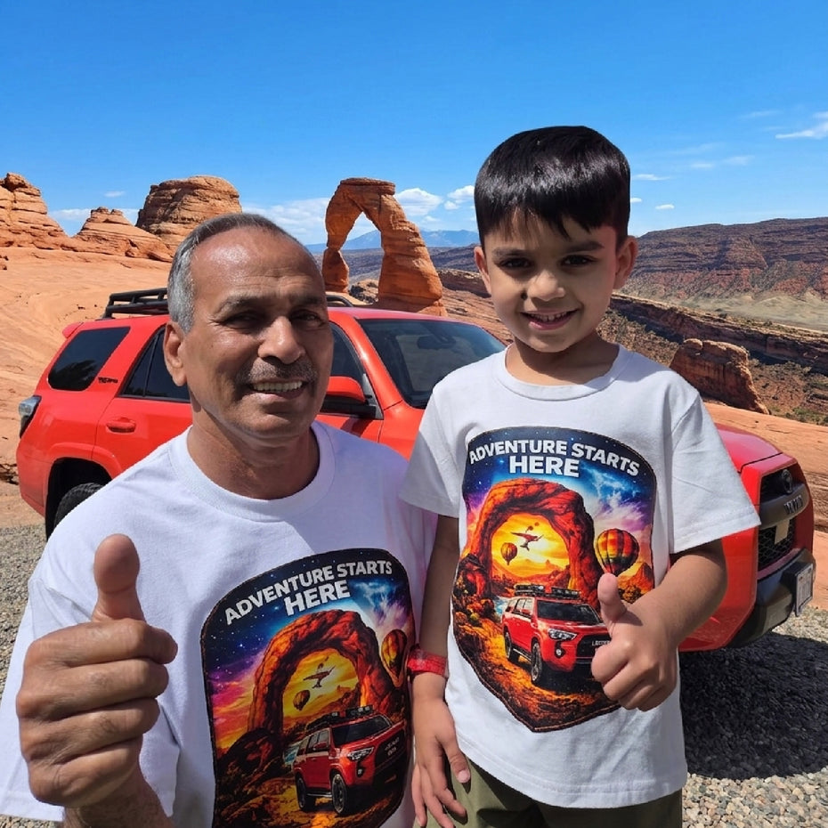 Man and child wearing 'Adventure Starts Here' t-shirts in a desert setting with a red SUV.